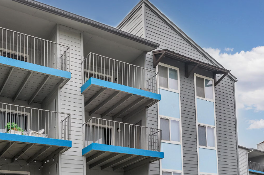 Apartment building with gray siding and blue balcony railings under a partly cloudy sky
