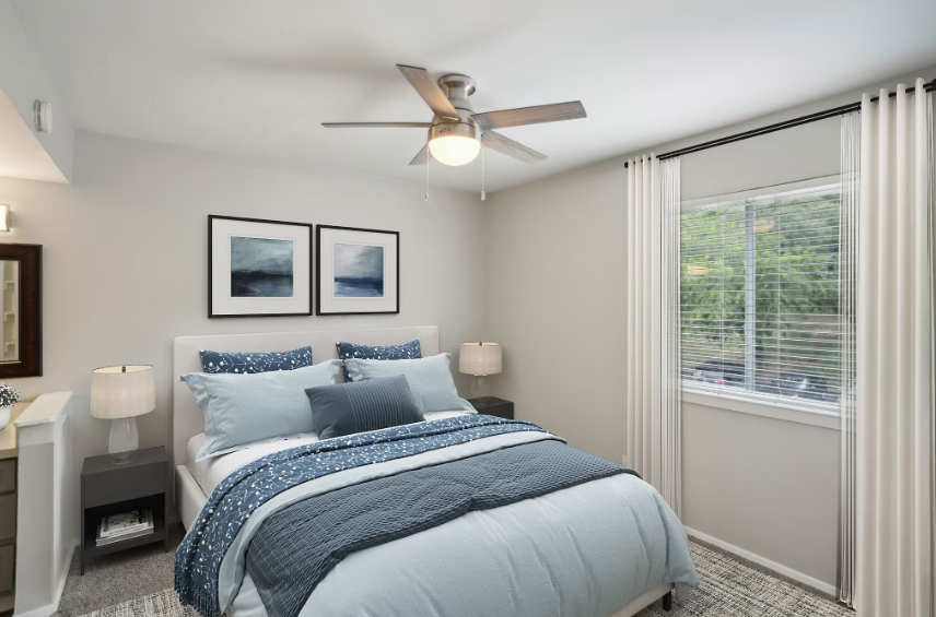 Bright bedroom with a bed, blue bedding, framed art, nightstands, and a window with blinds and vertical curtains