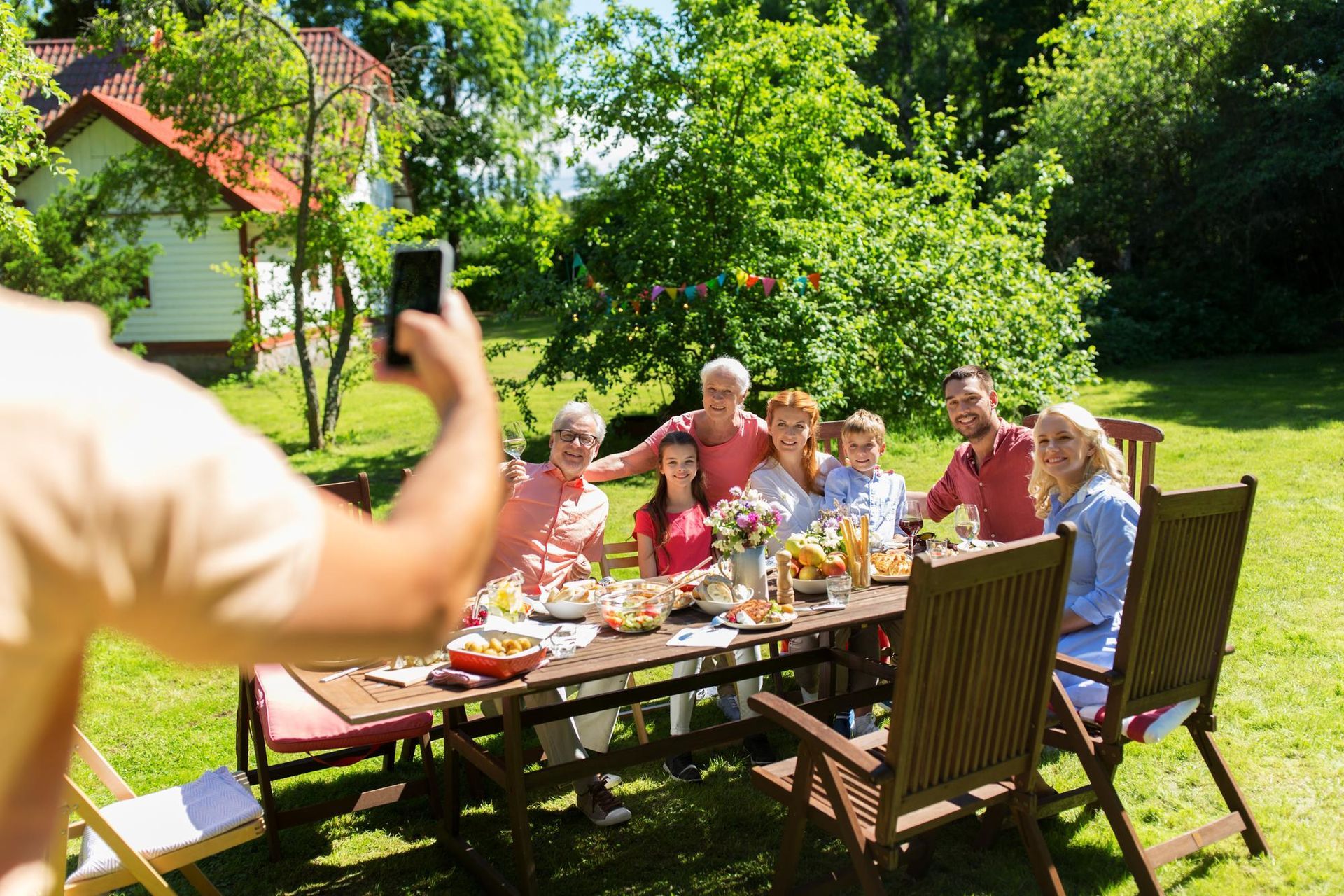 Friends sharing a meal outdoors at a garden table, with one person taking a photo on a sunny day