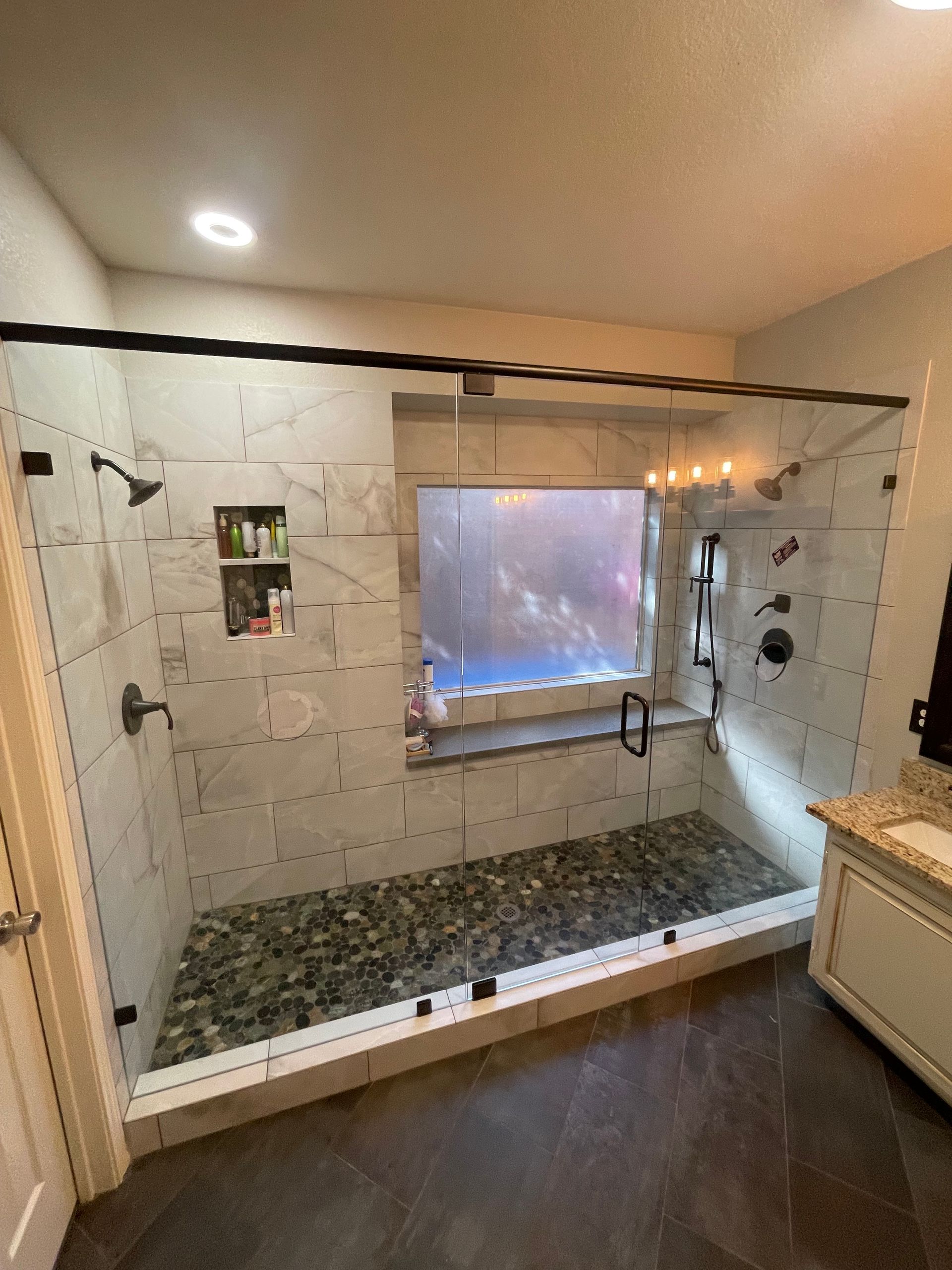 Spacious shower with glass doors and pebble floor. Marble tile walls, a window, and black fixtures are visible.