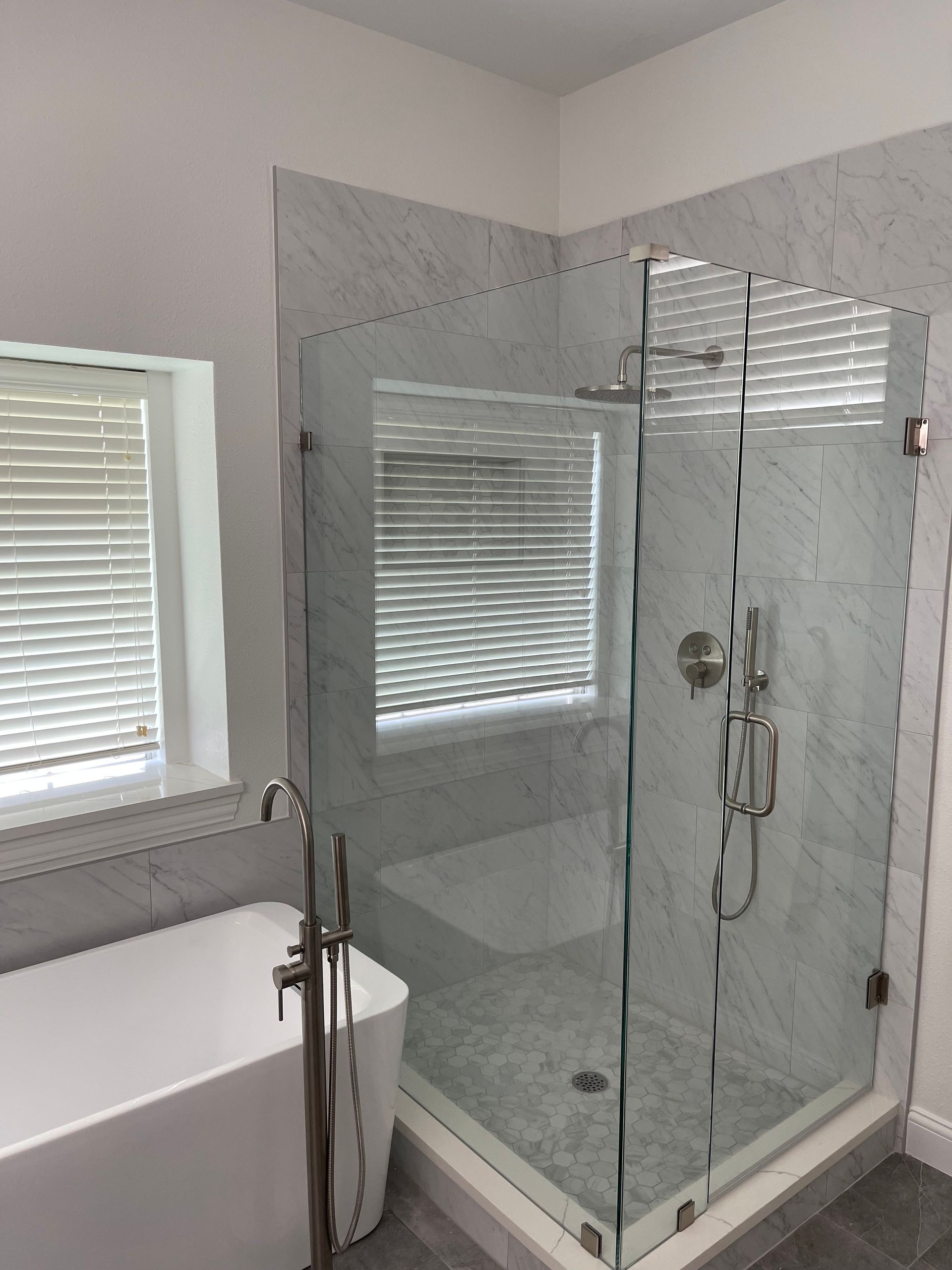 Modern bathroom with glass shower, white tub, marble tile, and window with blinds.