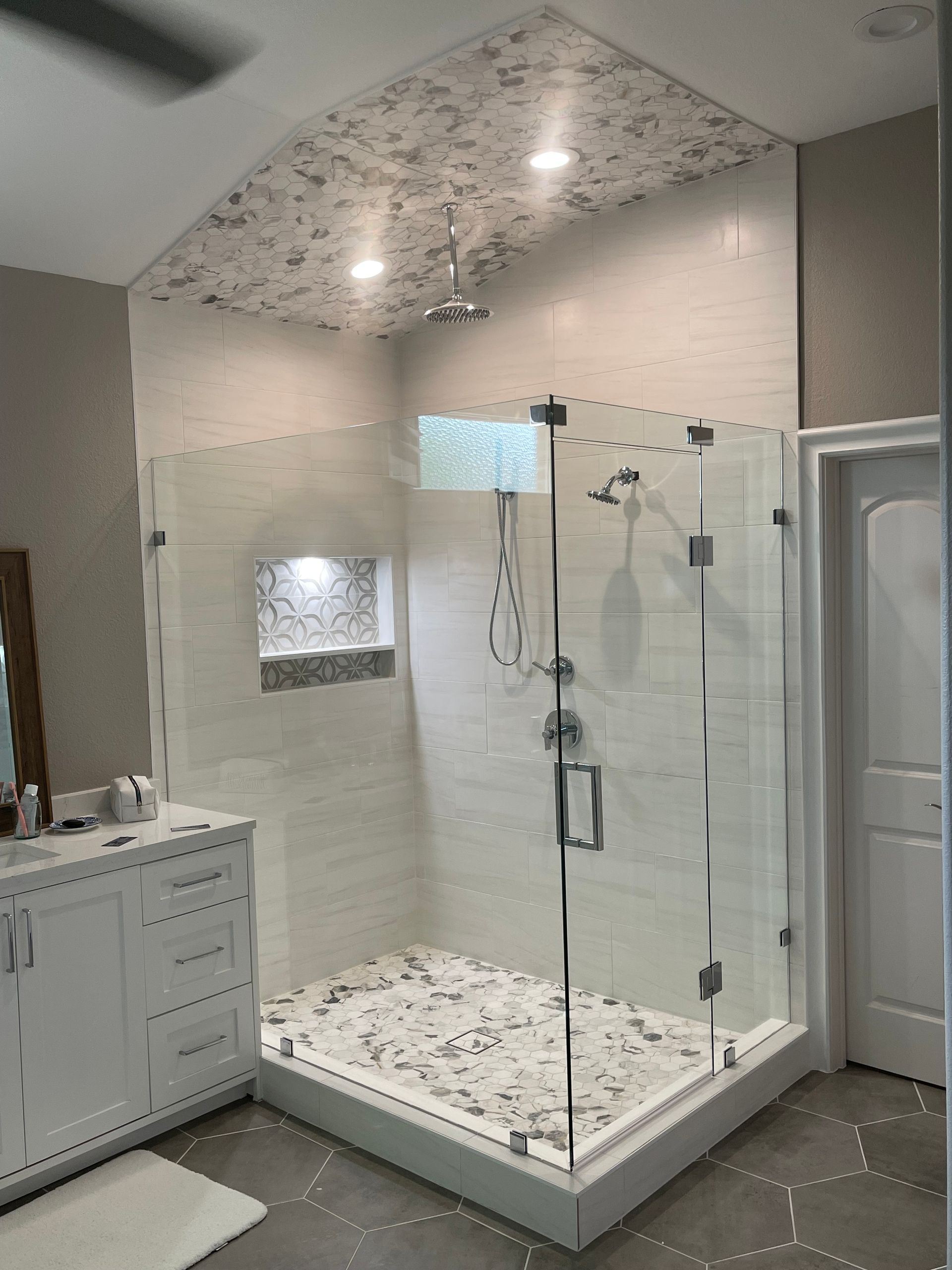 Modern bathroom with white tiled shower, glass doors, and patterned floor, alongside a white vanity.