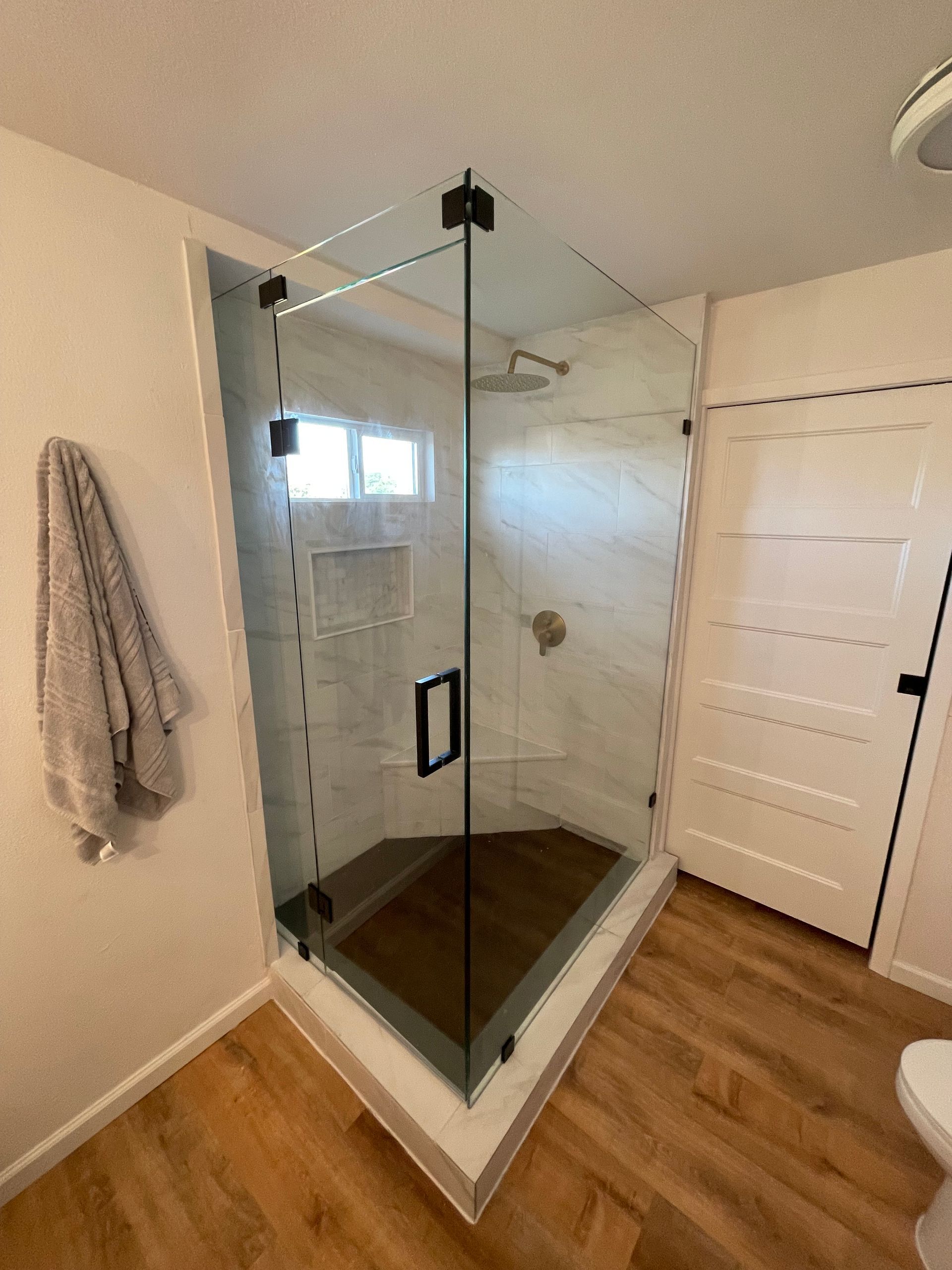 Corner shower with glass doors, black hardware, white tile, and wood-look flooring.
