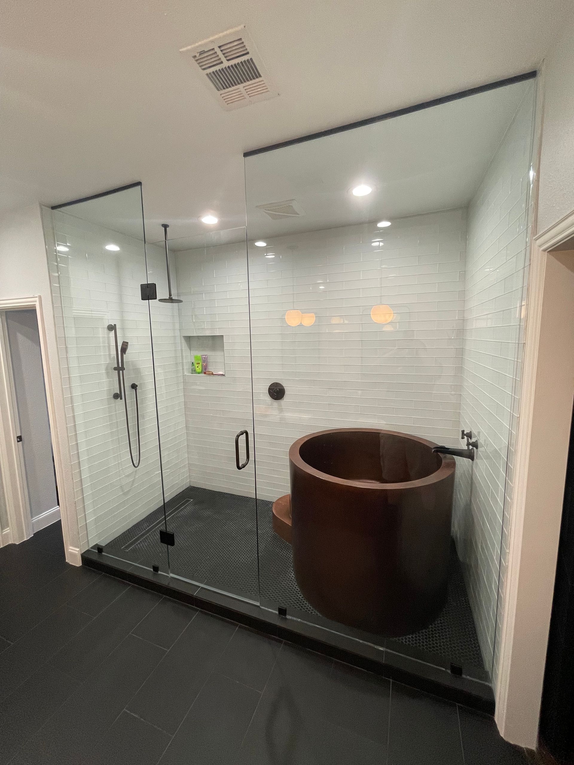 Modern shower with wooden soaking tub, glass walls, white tile. Black floor and fixtures.
