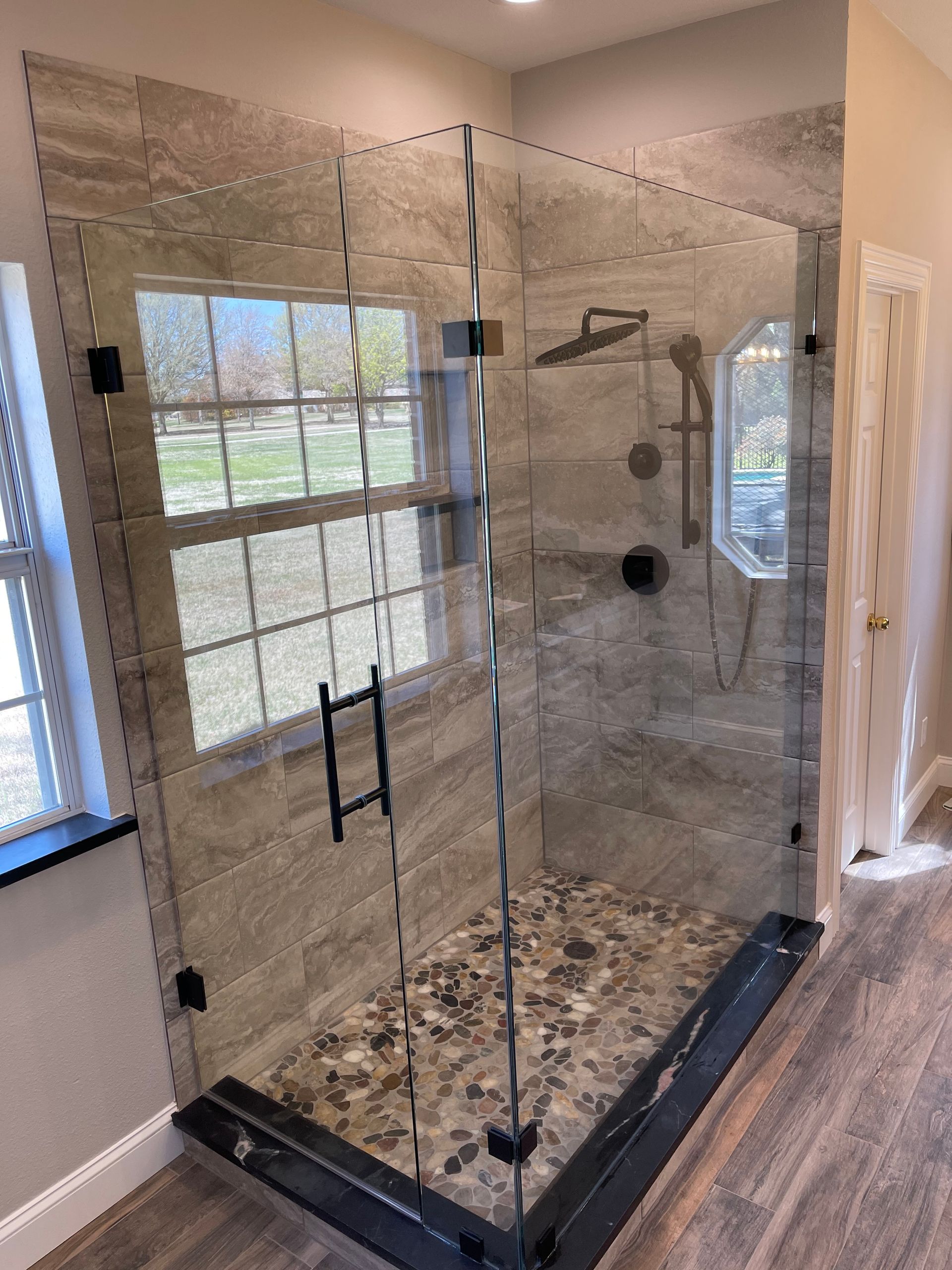 Glass shower enclosure with gray tile and pebble floor. Black fixtures, window view.