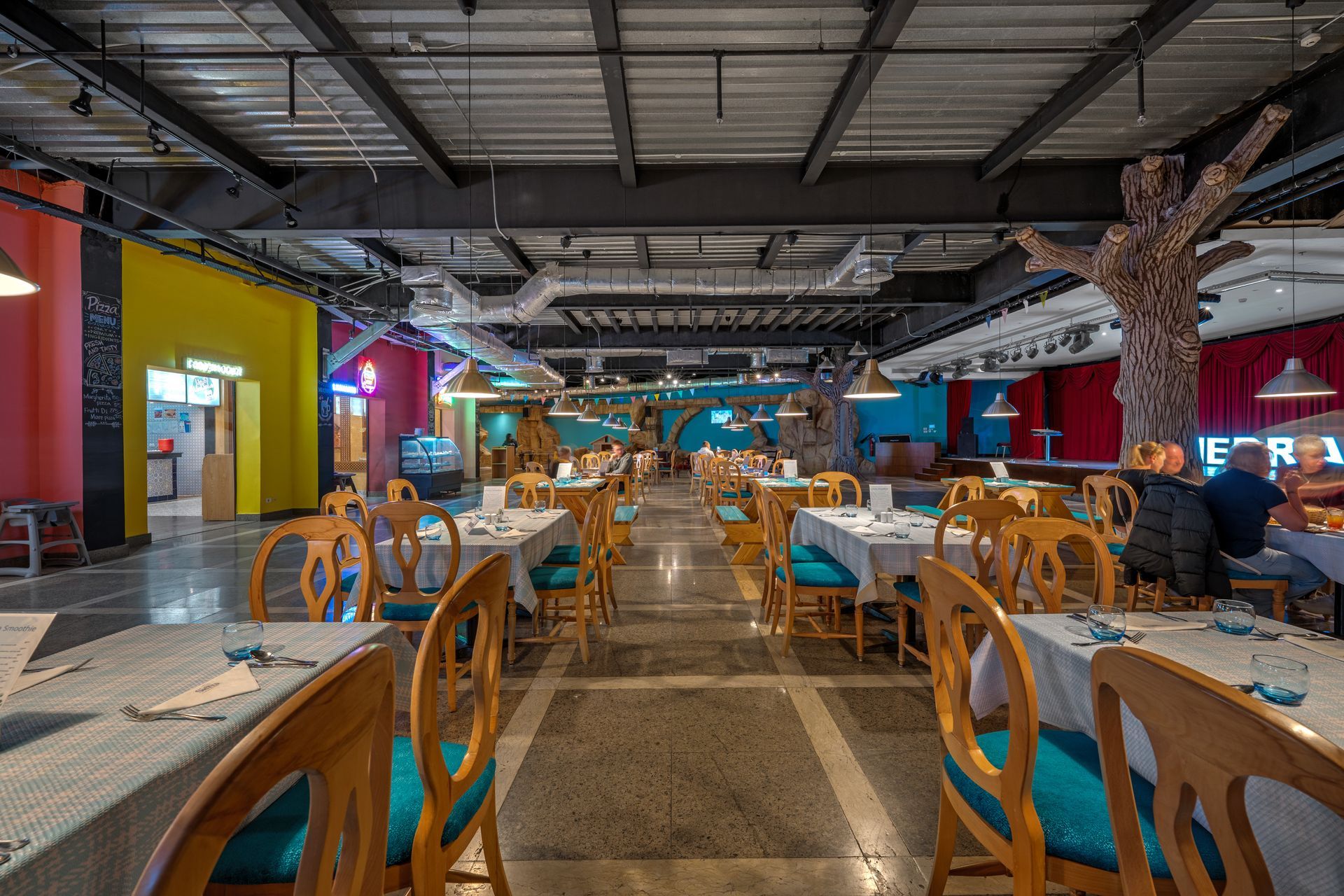 Restaurant interior with tables, chairs, colorful walls, and a decorative tree.