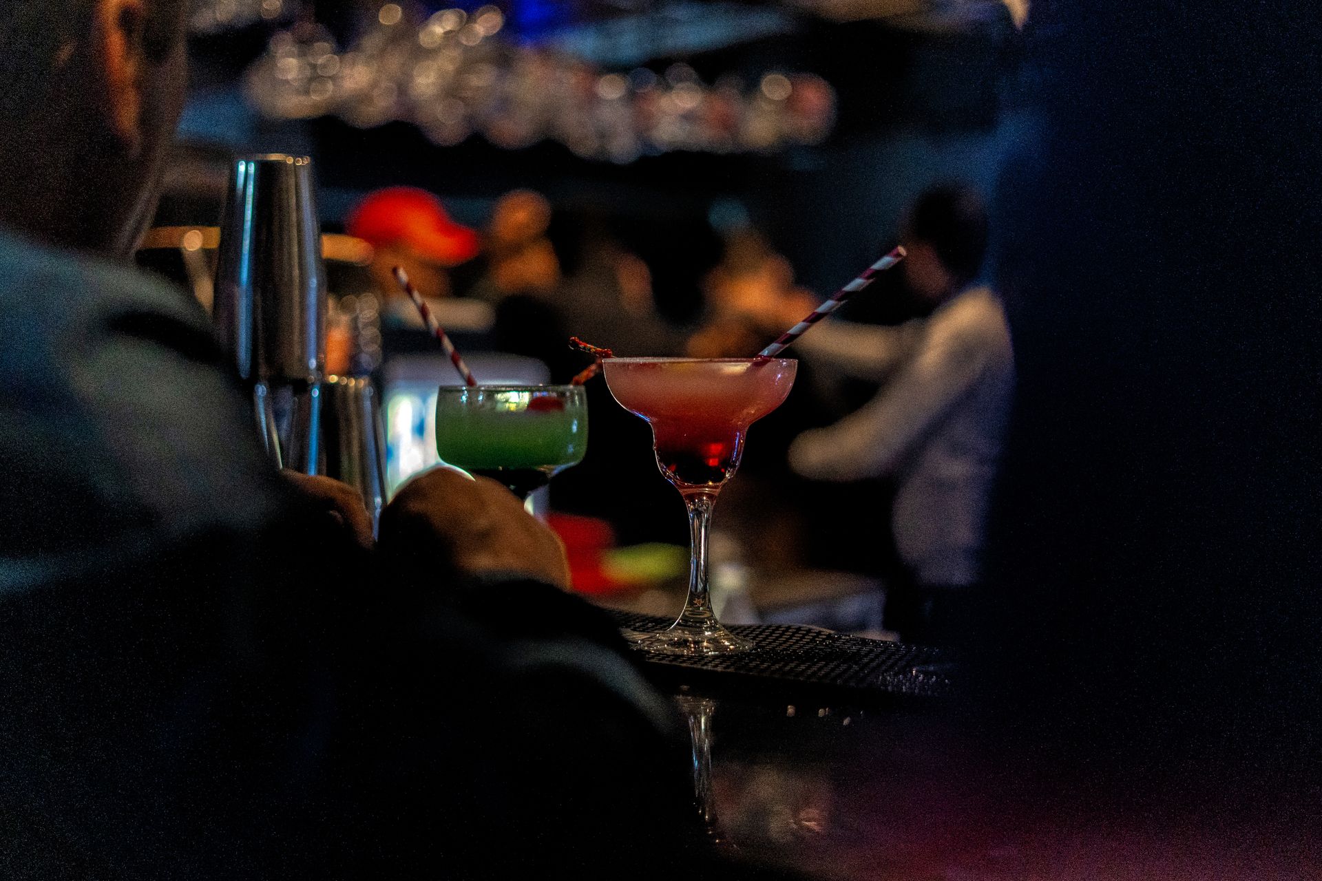 Two cocktails on a tray in a dark bar, with a bartender in the background.
