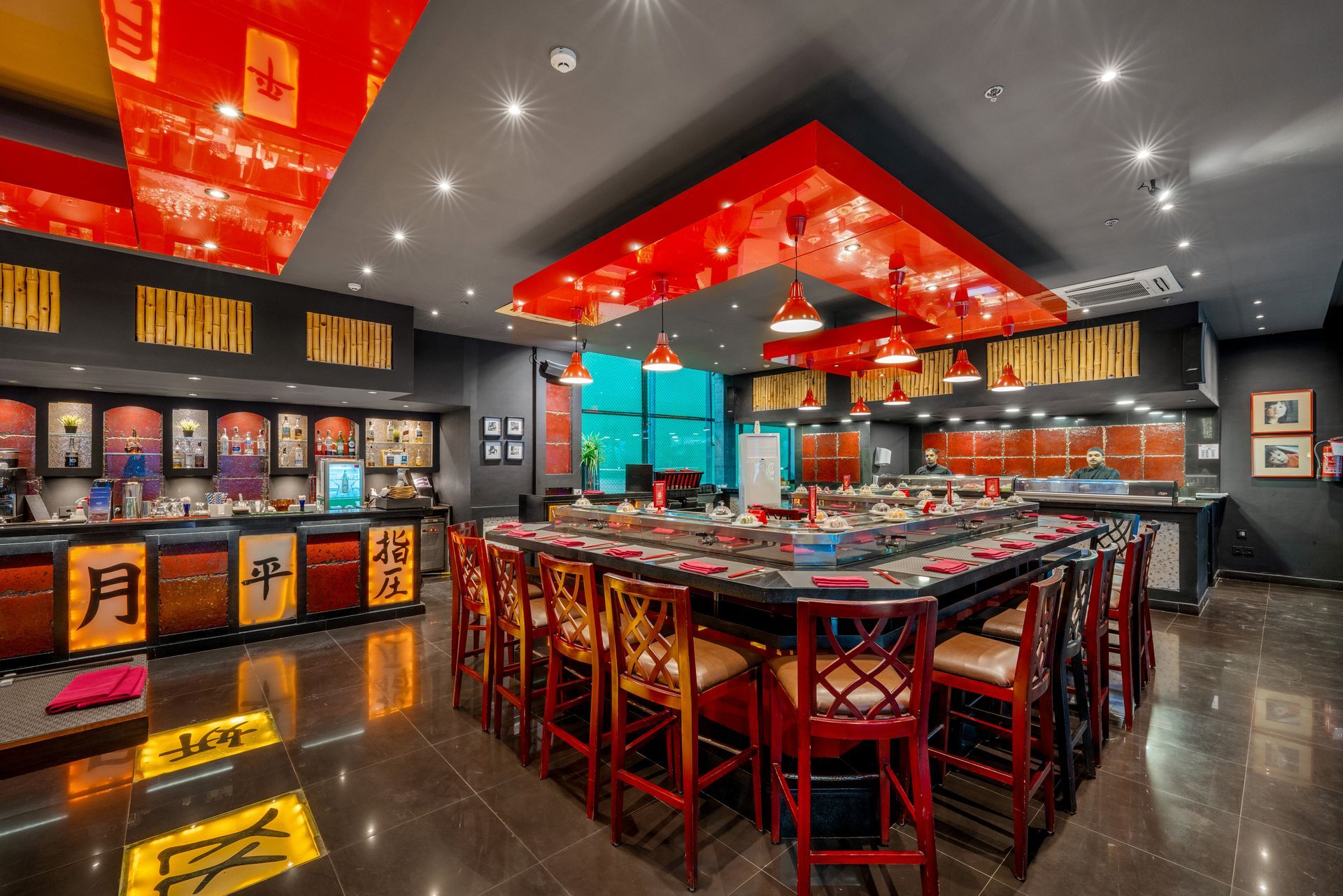Interior of a brightly lit Japanese restaurant. Red accents on ceiling, chairs, and walls. Sushi bar.