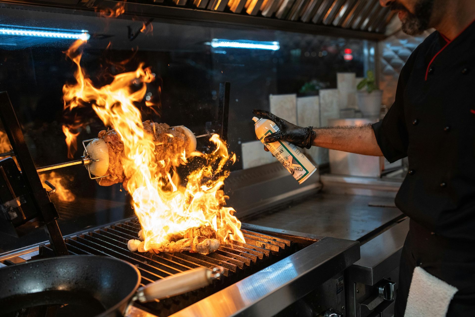 Chef in black uniform spraying flames on grill, creating large fire in kitchen.