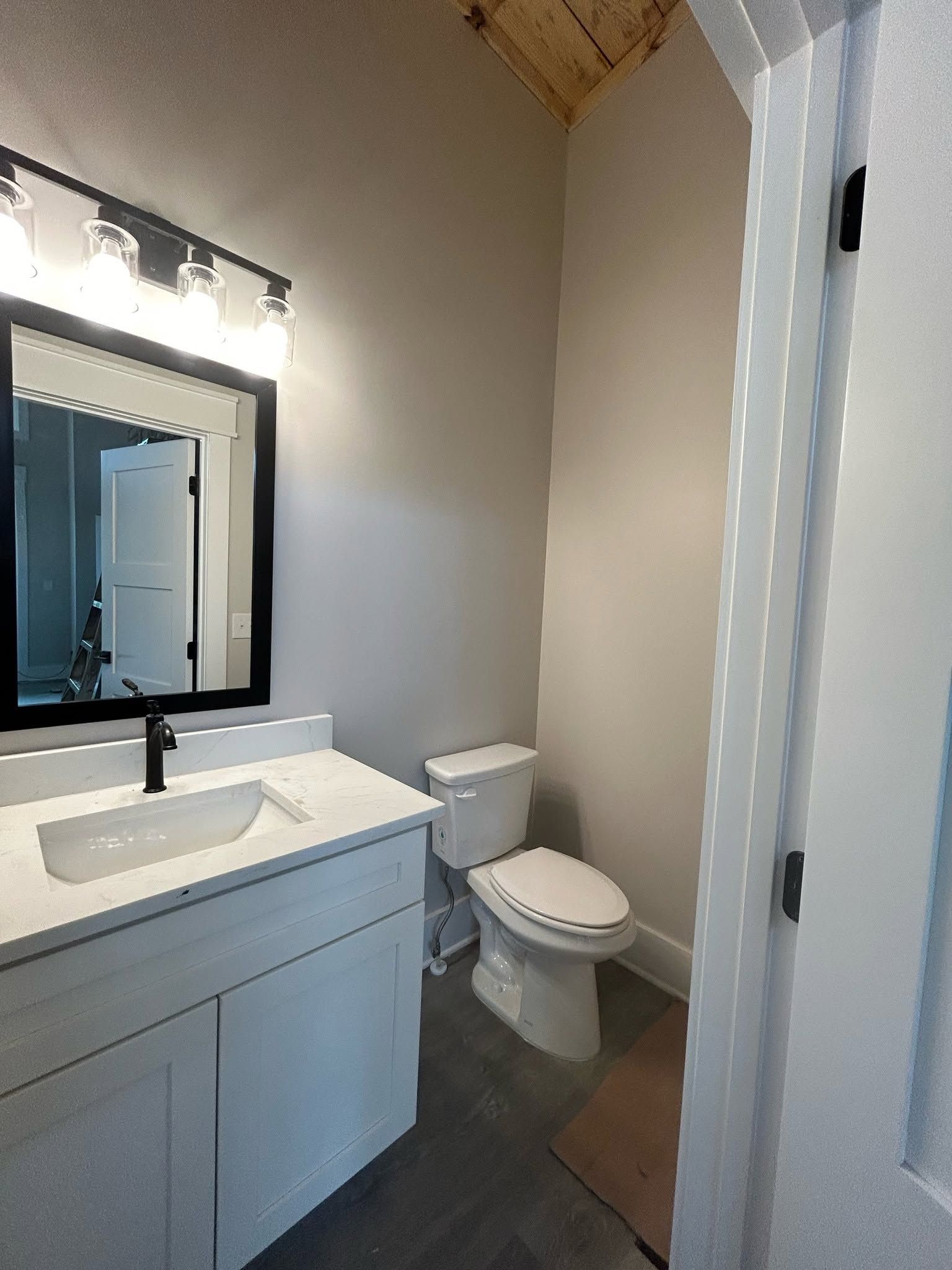 Small bathroom with white vanity, mirror, toilet, and gray walls under bright vanity lights