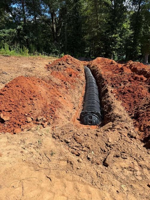 Black corrugated pipe laid in a red dirt trench at a wooded construction site