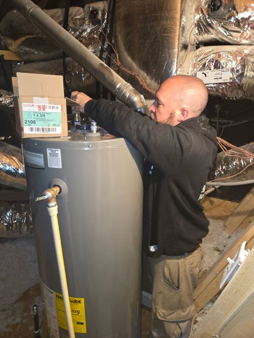 Worker inspecting a water heater in a basement with exposed ducts and pipes