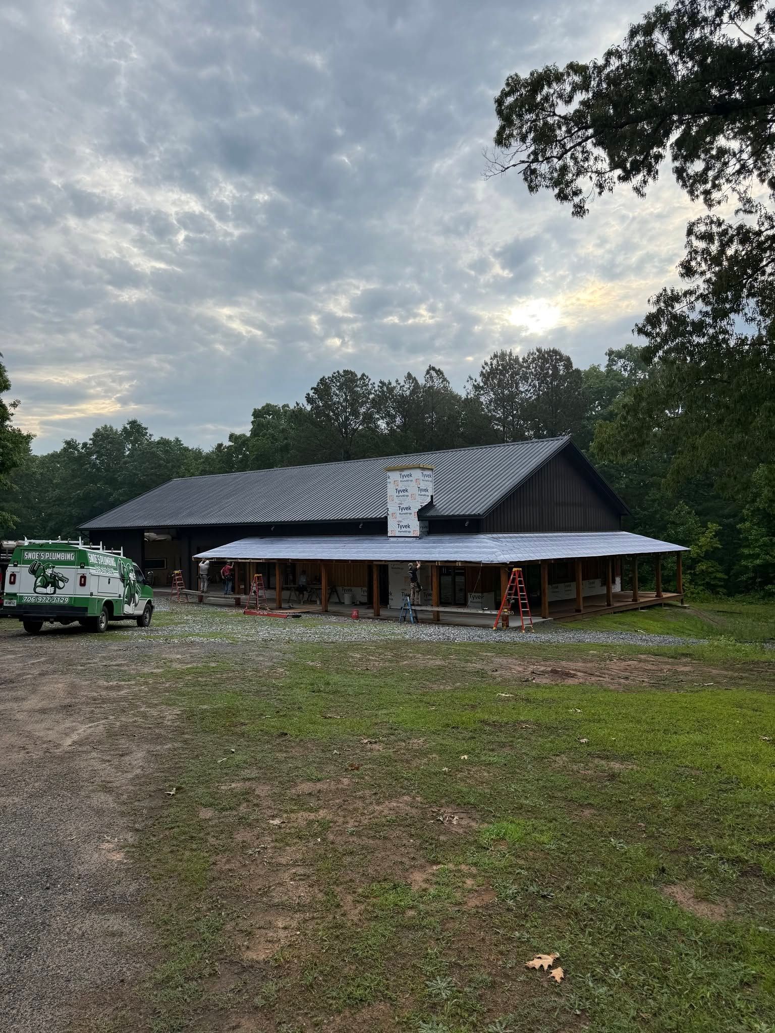 Long building with a striped roof in a wooded clearing at dusk, with trucks parked nearby