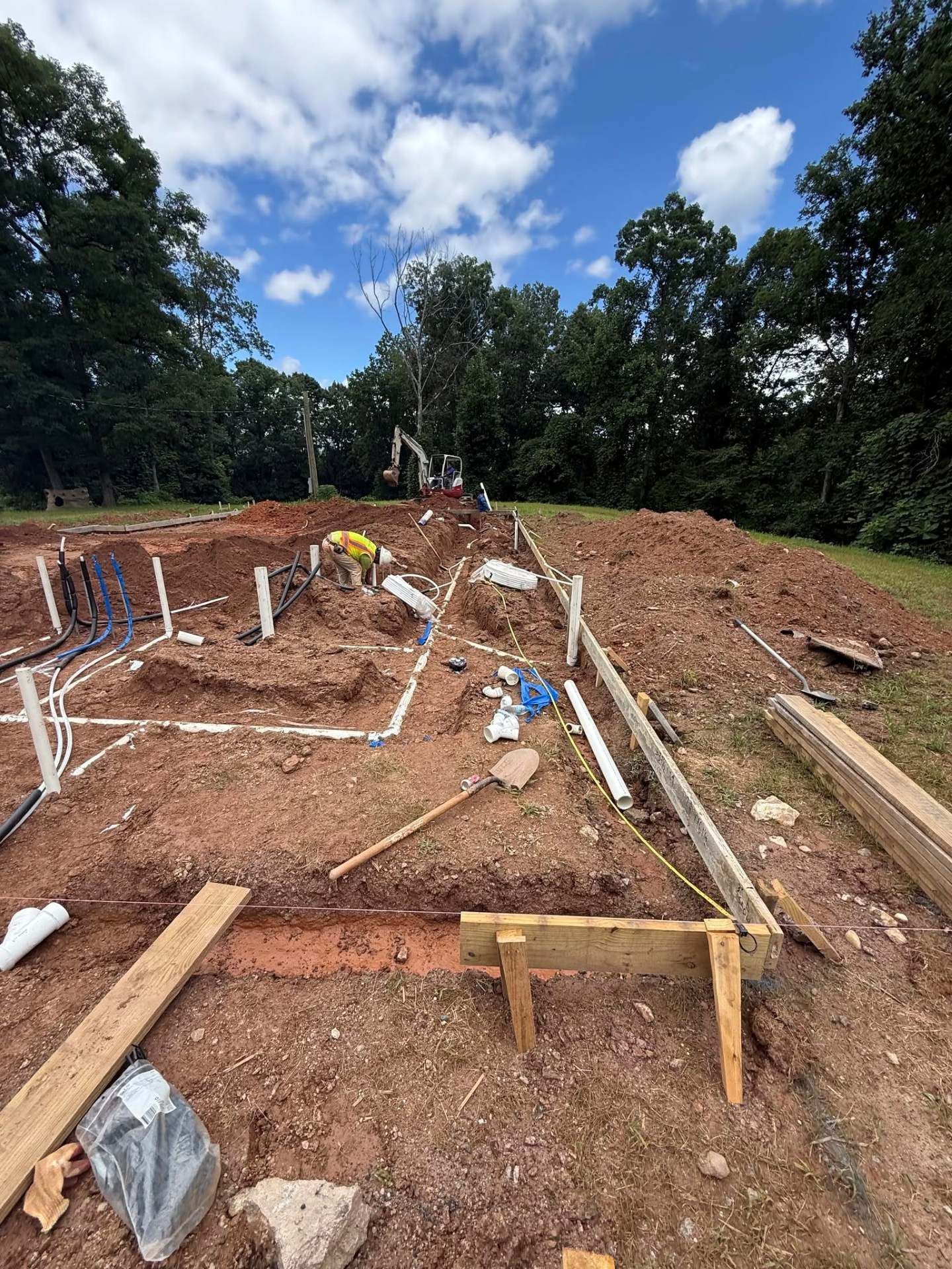 Construction site with foundation trenches, lumber framing, and dirt piles under a cloudy blue sky