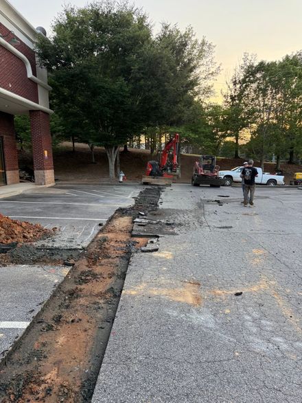 Construction equipment and workers repairing a cracked parking lot beside a brick building at dusk