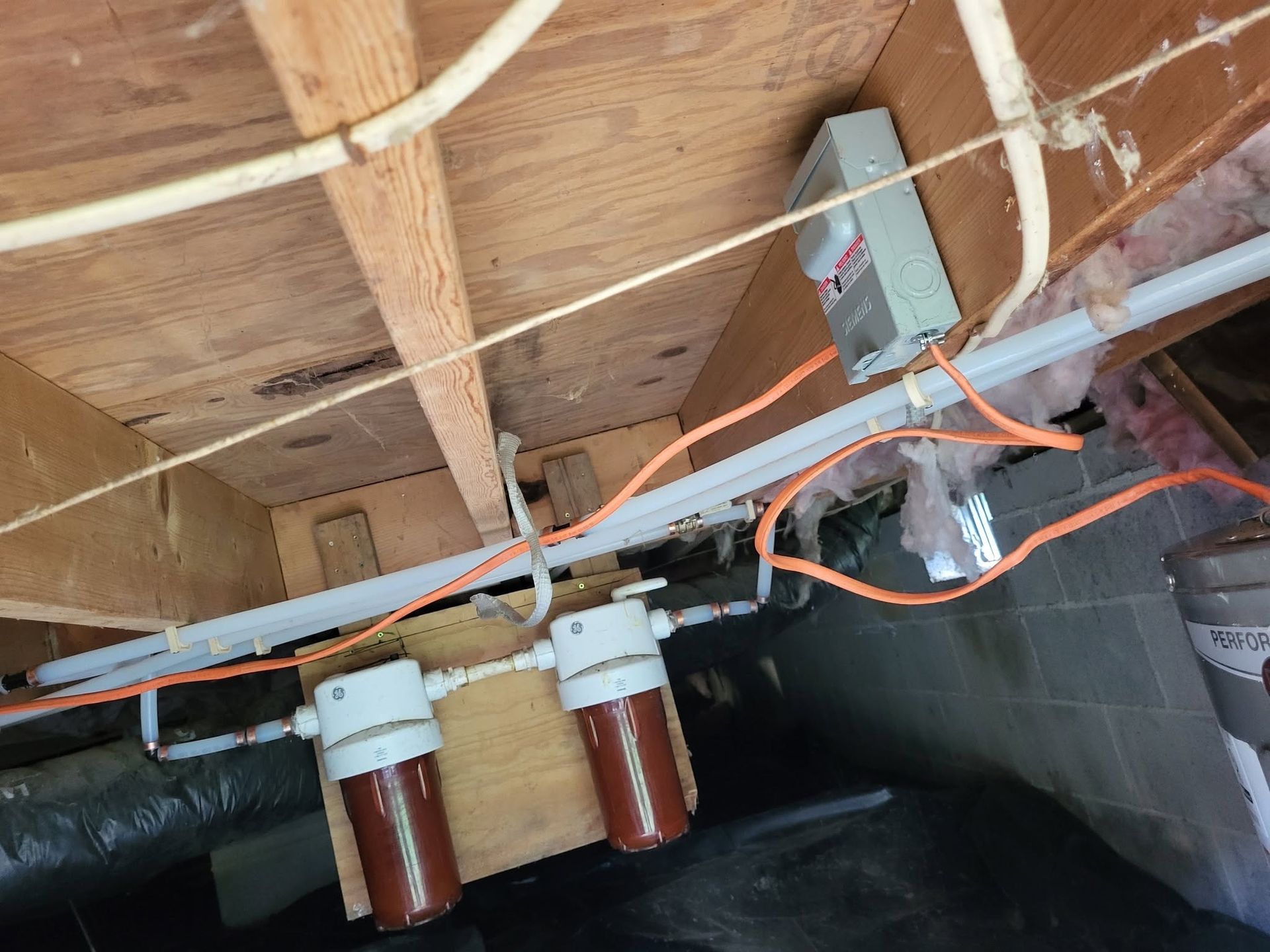 Exposed plumbing and electrical wiring under a floor joist in an unfinished crawlspace.
