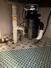 Under-sink plumbing with a black garbage disposal, white pipes, and a patterned floor mat