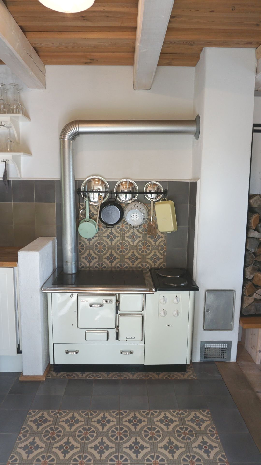 Weißer Küchenherd mit Metallrohren und hängenden Töpfen. Gefliester Spritzschutz, Holzdecke und gemusterter Boden.