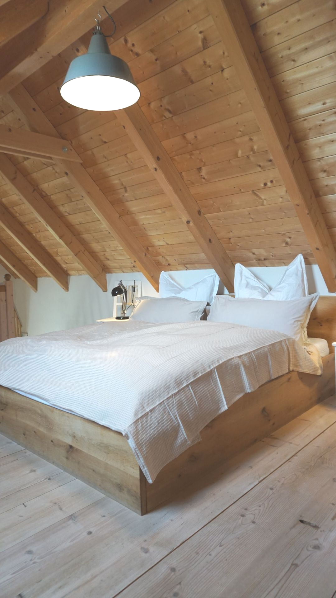 Schlafzimmer mit Holzelementen: Dachschräge, Bett mit weißer Bettwäsche, Pendelleuchte darüber.