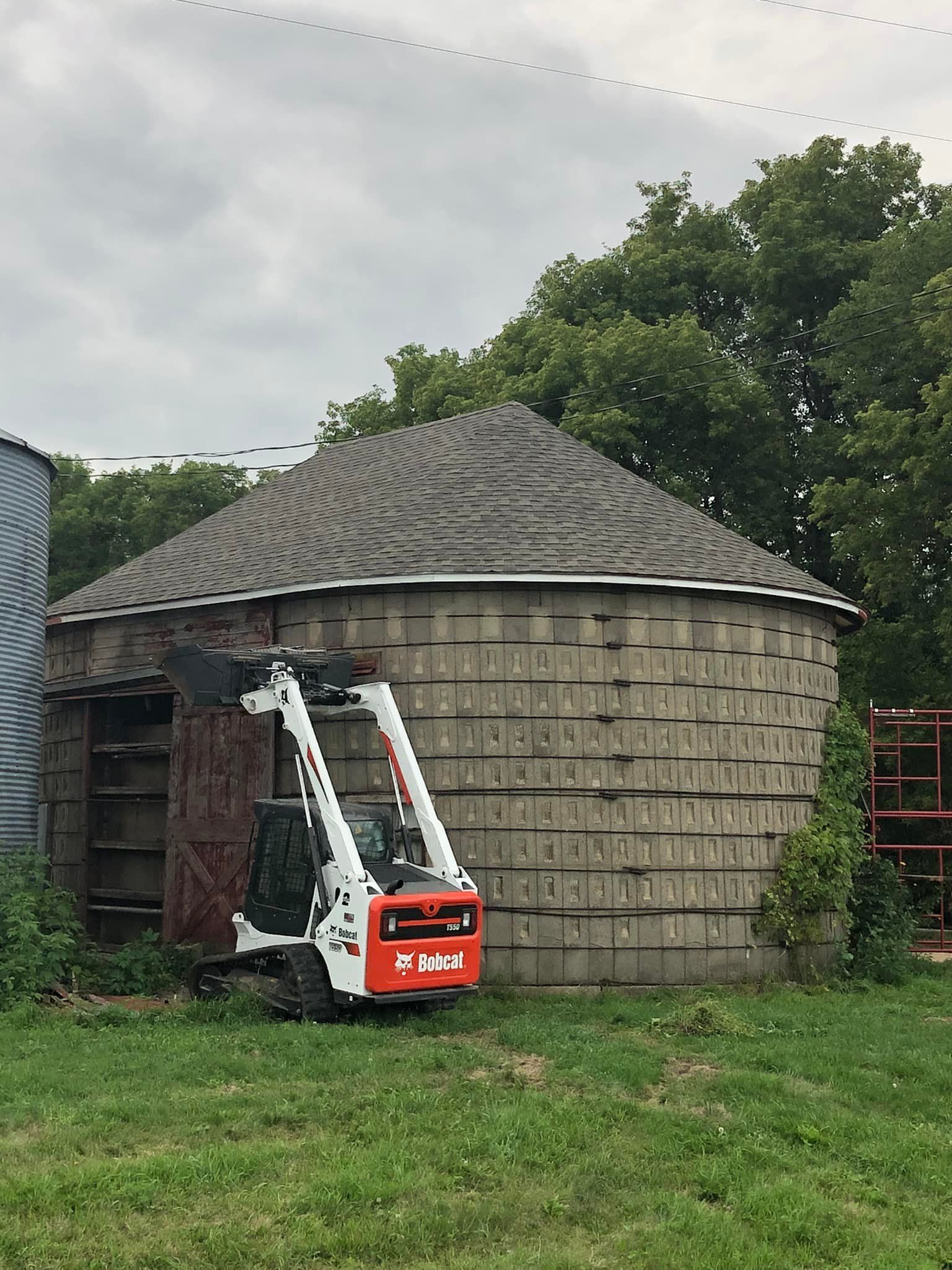 Bobcat skid-steer loader parked in front of a weathered, cylindrical silo. Overcast day.