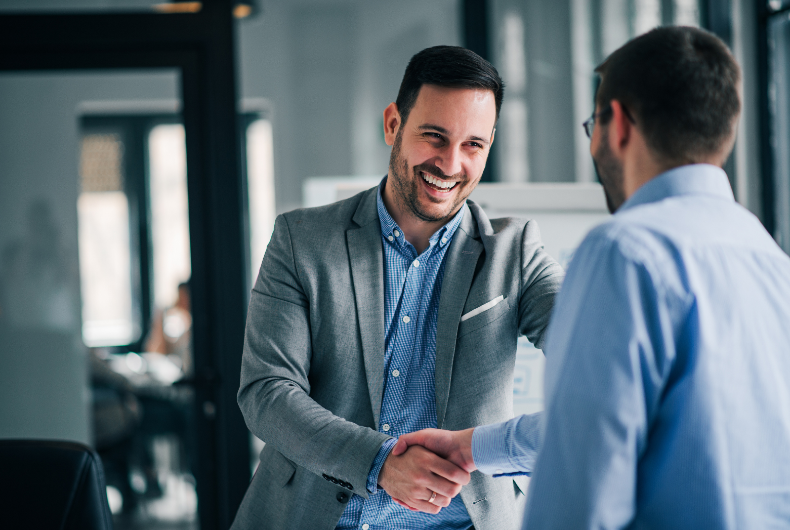 Two professionals in office attire smiling and shaking hands in a professional setting.