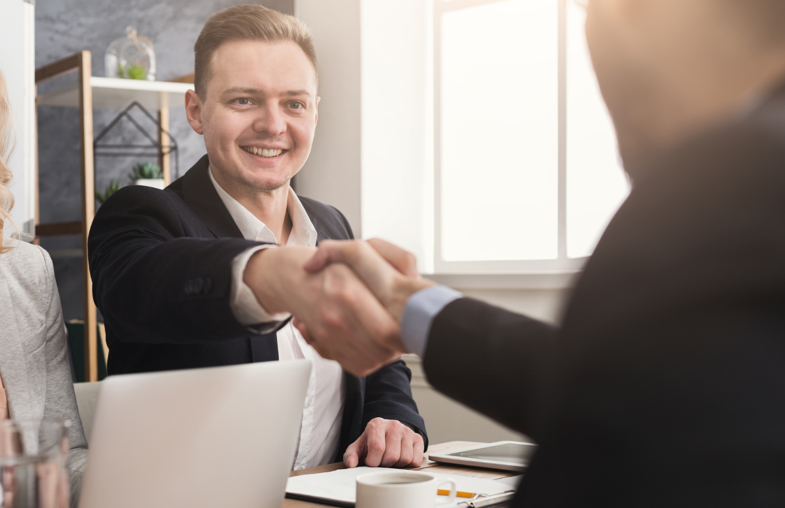 Two professionals in business attire shaking hands across an office desk, smiling during a meeting.
