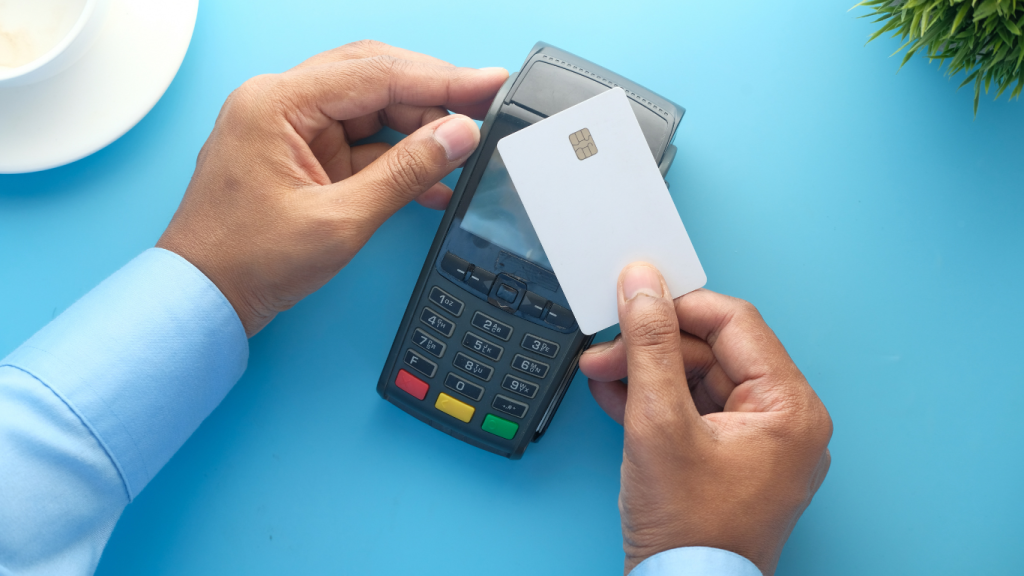 A person in a light blue shirt holds a blank white credit card over a payment terminal on a solid blue surface.