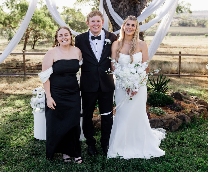 Bride and groom with a woman, posing outdoors. Bride holds bouquet, all smiling.