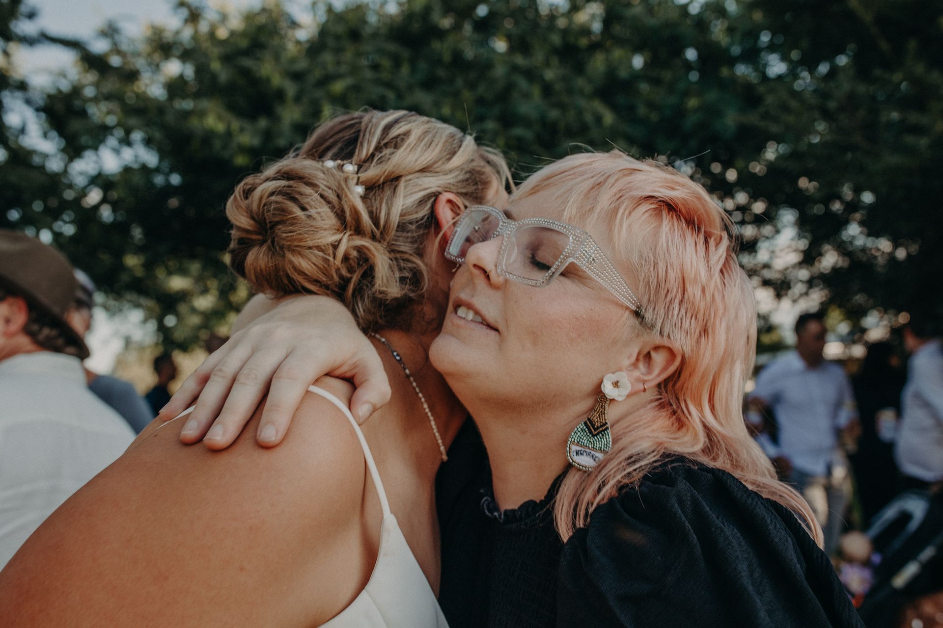Two women embrace outdoors; one with pink hair and bedazzled glasses, the other in a white dress with a bun.