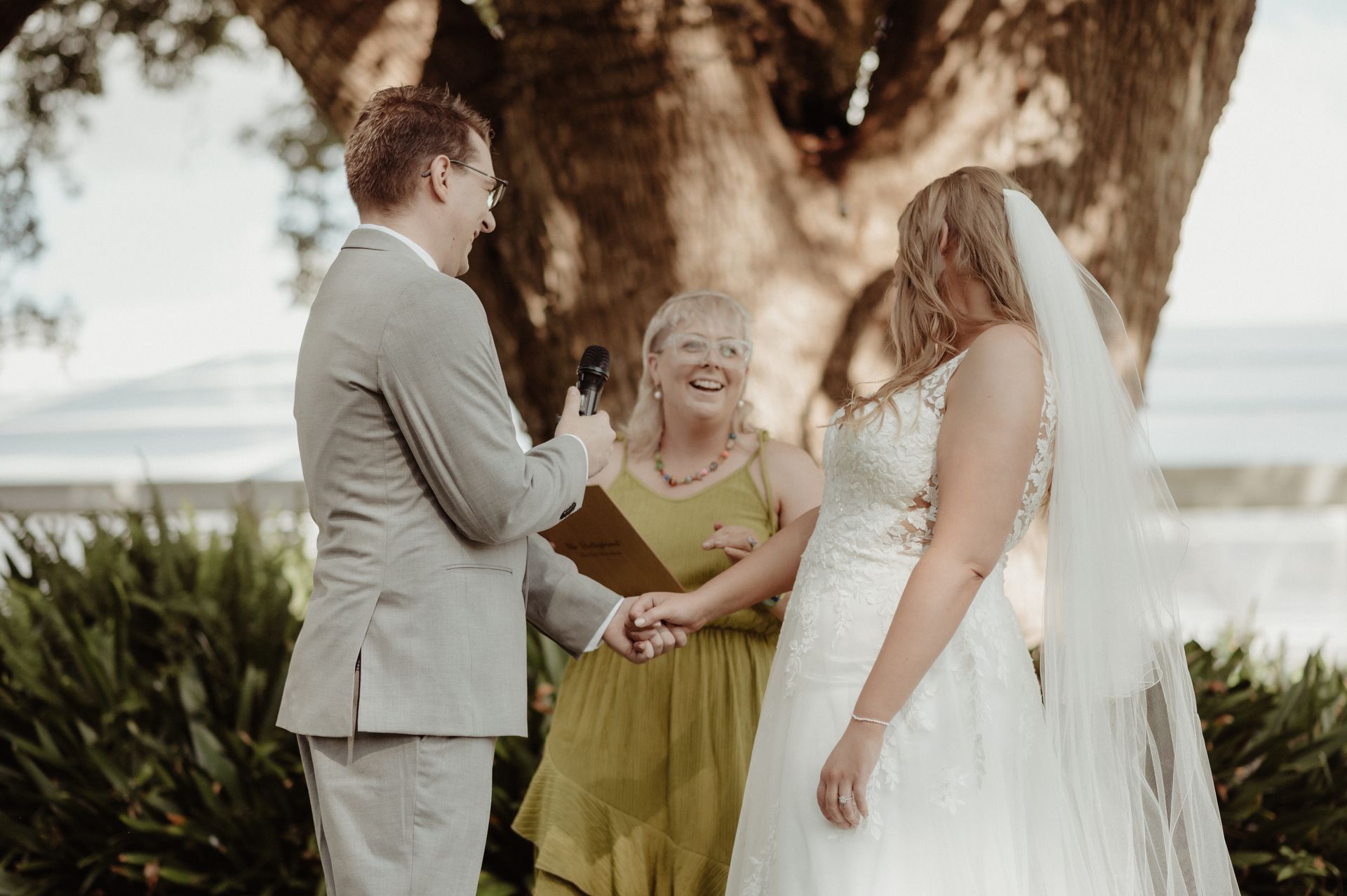Wedding couple and officiant under a large tree, smiling outdoors in soft sunlight.
