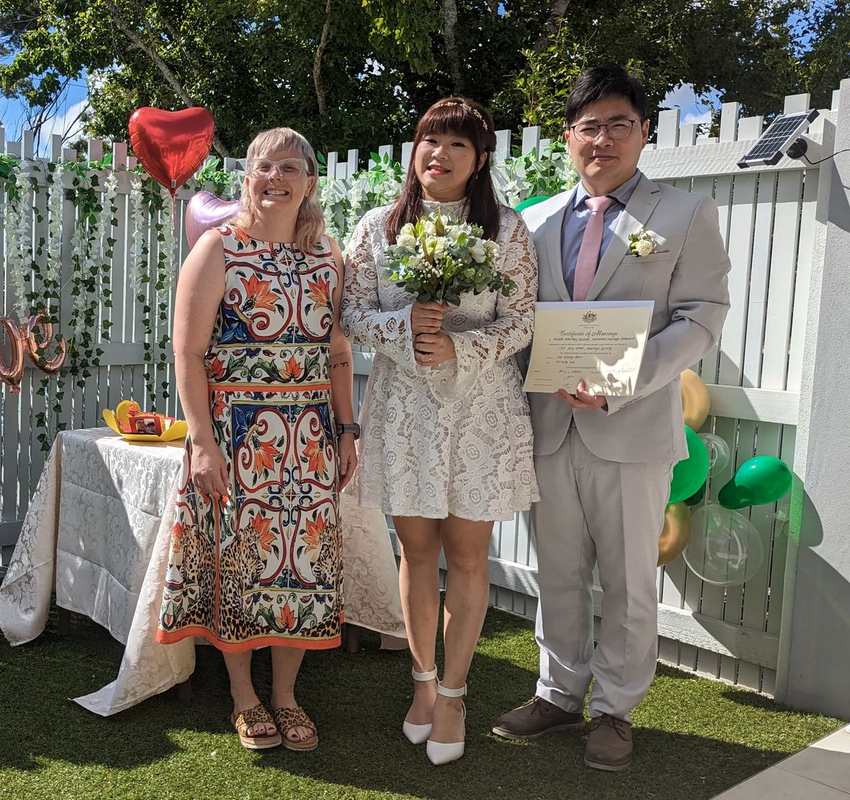 A wedding ceremony outdoors: three people pose, including a bride in white and a groom in a light suit.