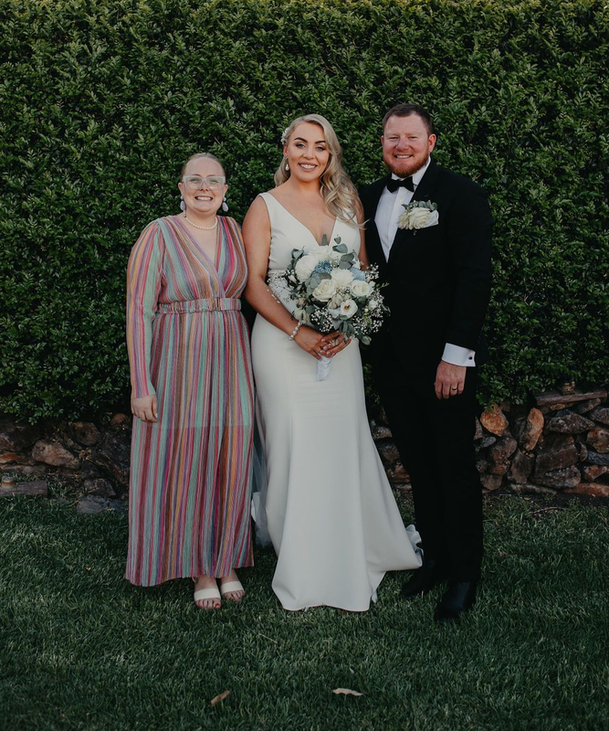 Bride and groom pose with another person. They stand in front of a green hedge. The bride holds a bouquet.