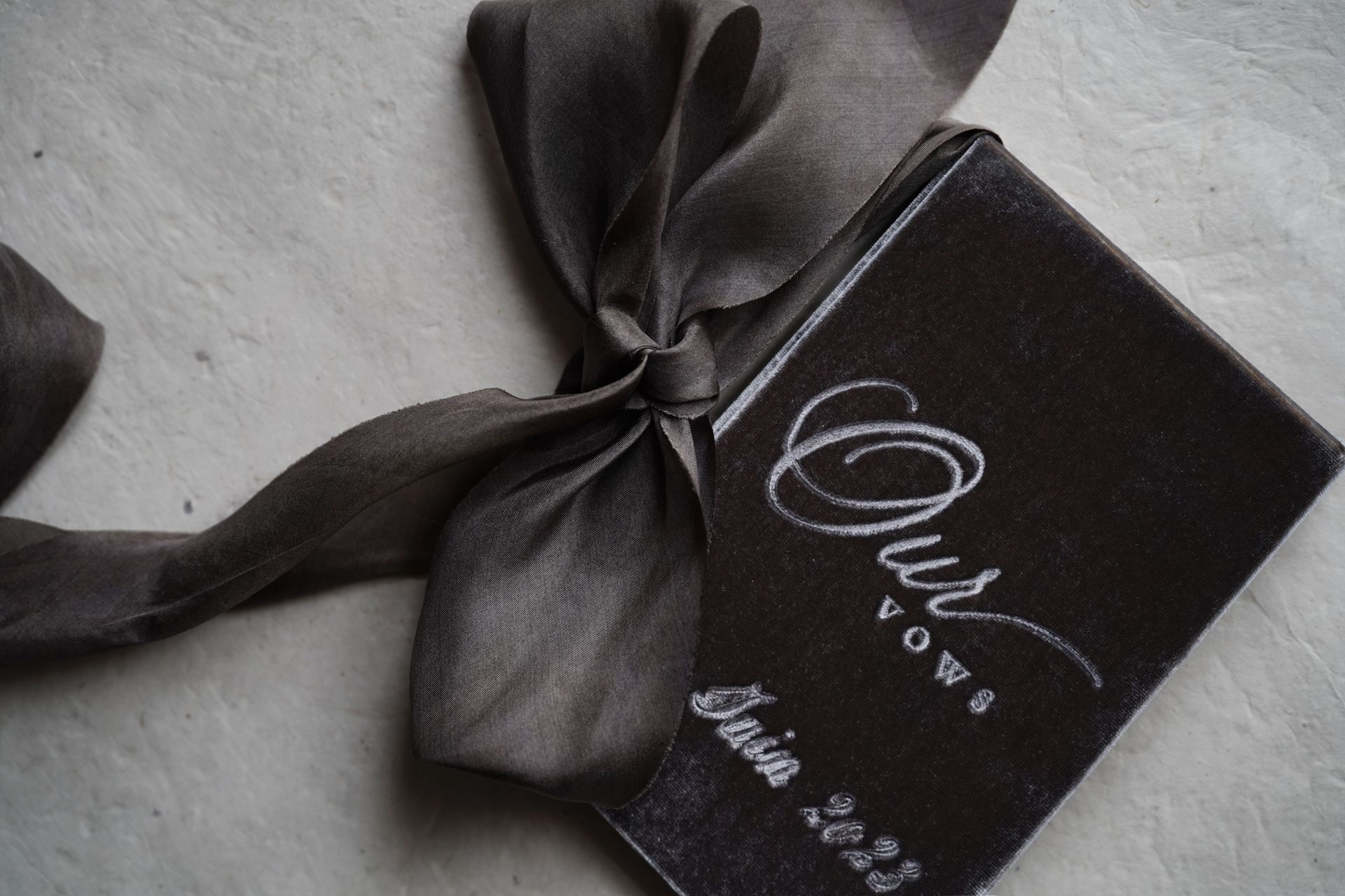 Black gift box with satin ribbon and white script logo on a grey surface