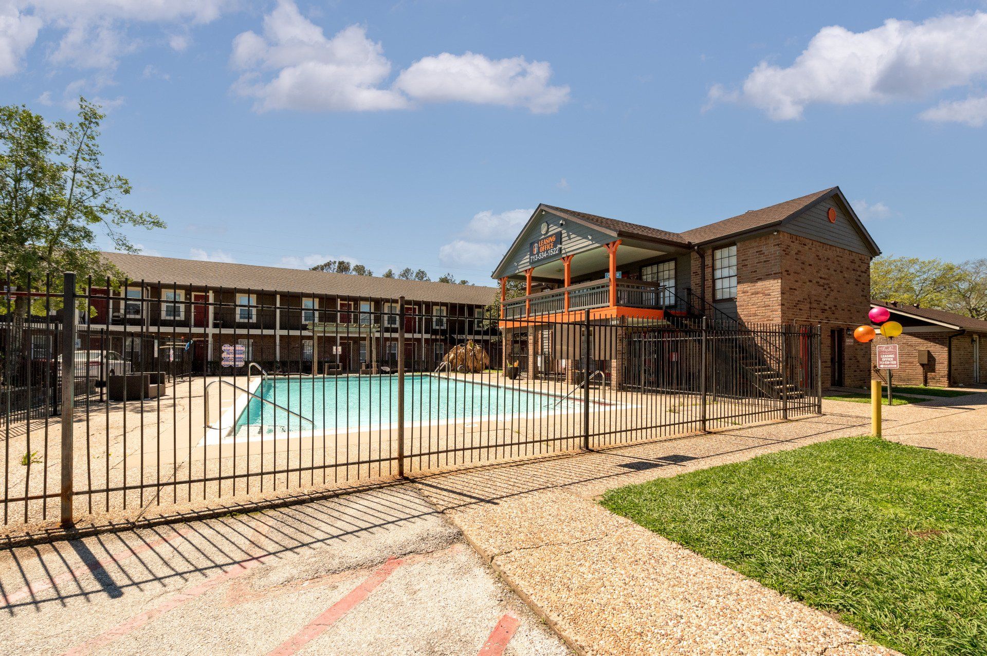 A fenced swimming pool area in front of two-story brick apartment building with a sign for leasing.