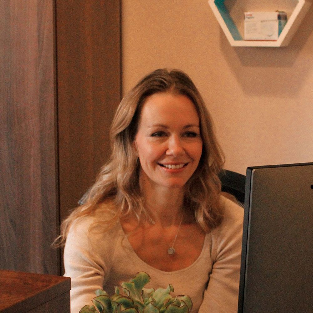 Woman smiling, sitting at a desk with a computer, light-colored sweater, office setting.