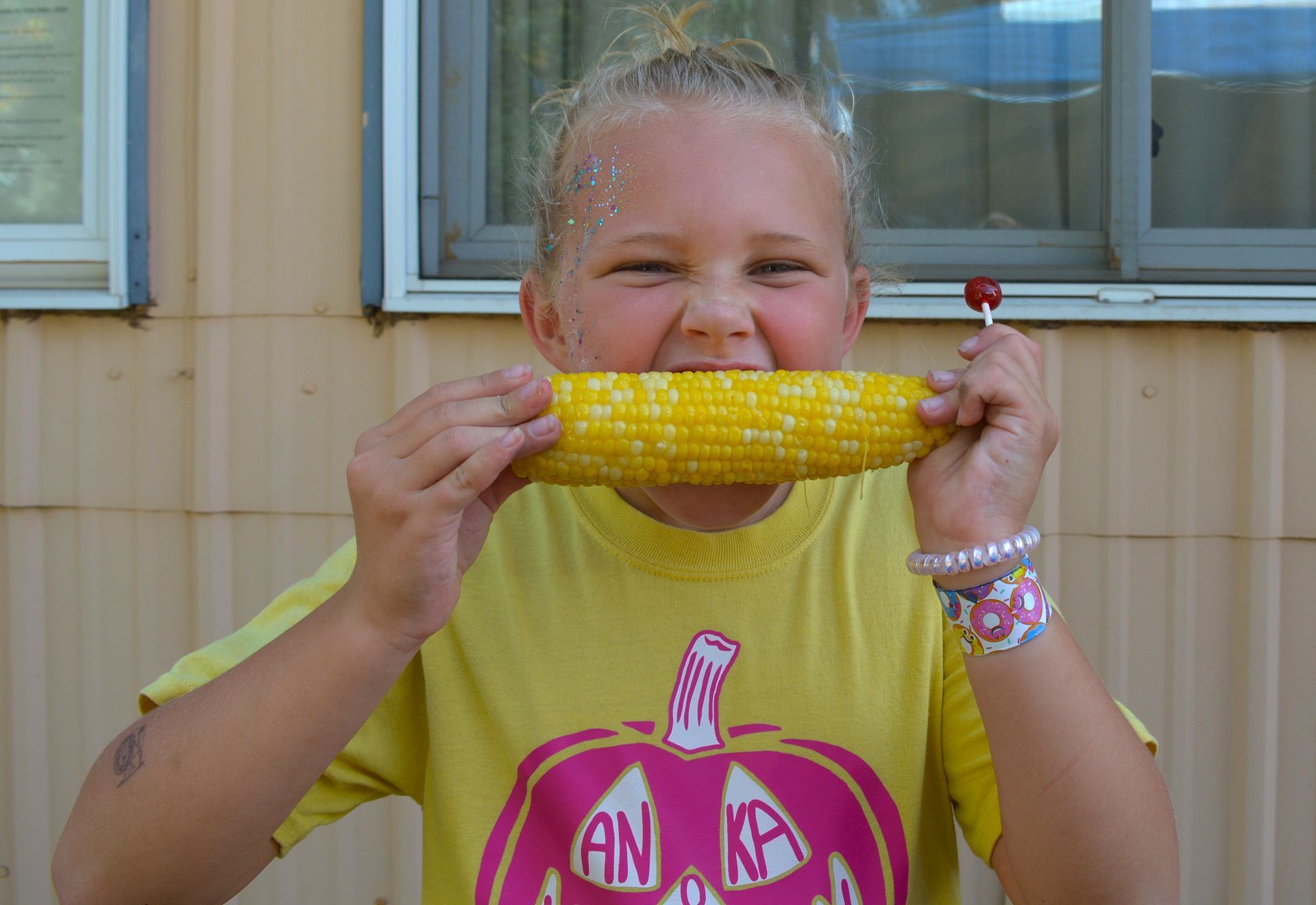 Person holding a corn cob to their mouth and smiling, wearing a yellow shirt outdoors.