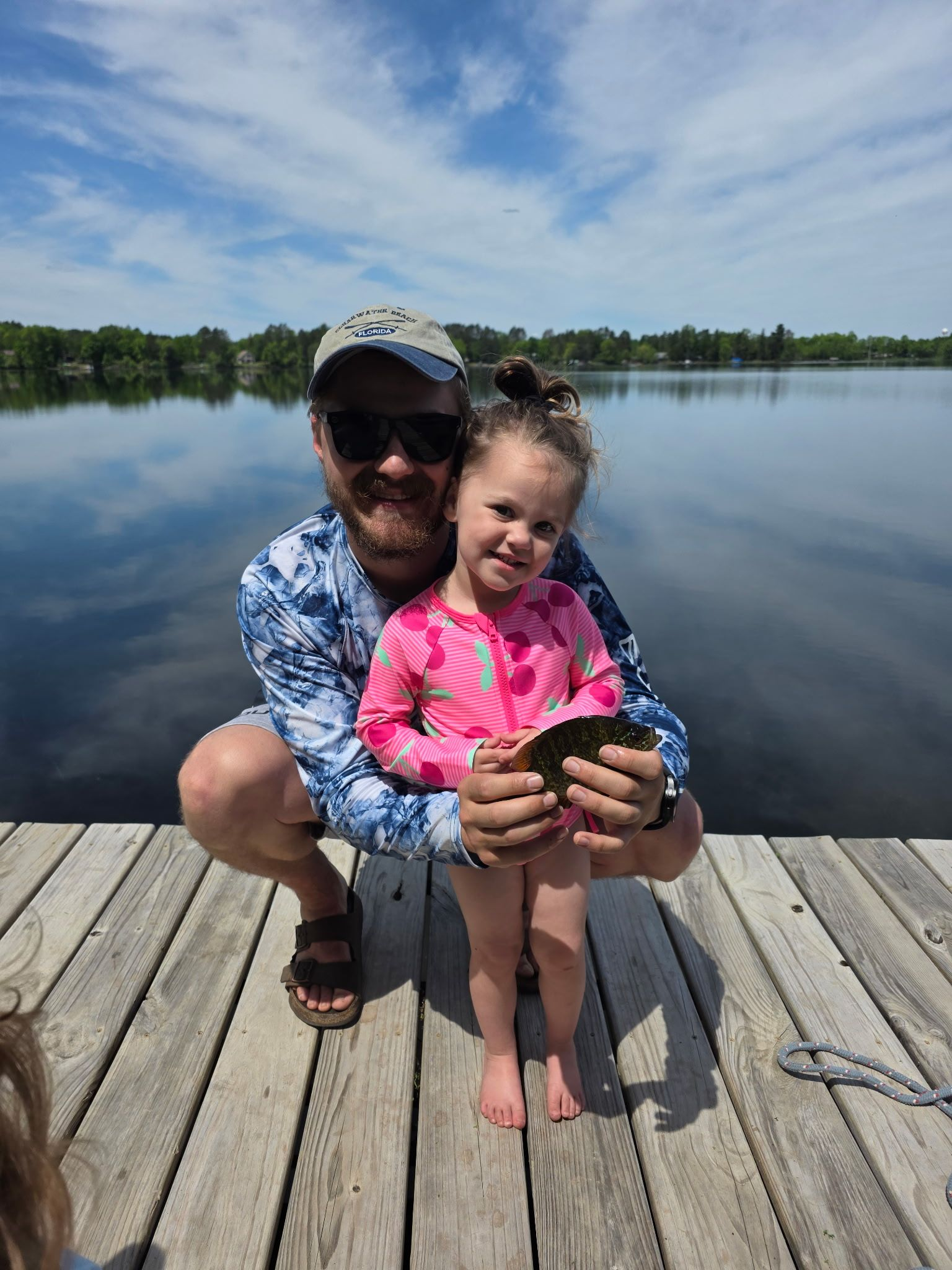 Two people sitting on a dock by a calm lake under a blue sky, smiling and holding a sunfish