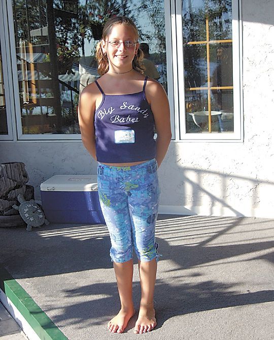 Person standing barefoot outdoors in a purple tank top and blue patterned leggings, smiling near a building.