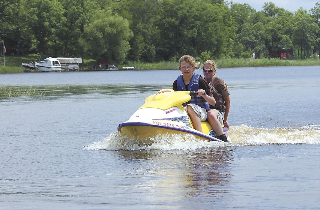 Two people ride a yellow jet ski across a lake with trees and a camper in the background