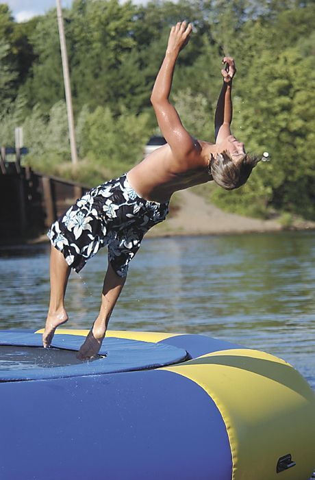 Person mid-backflip on a blue inflatable over water, with trees in the background