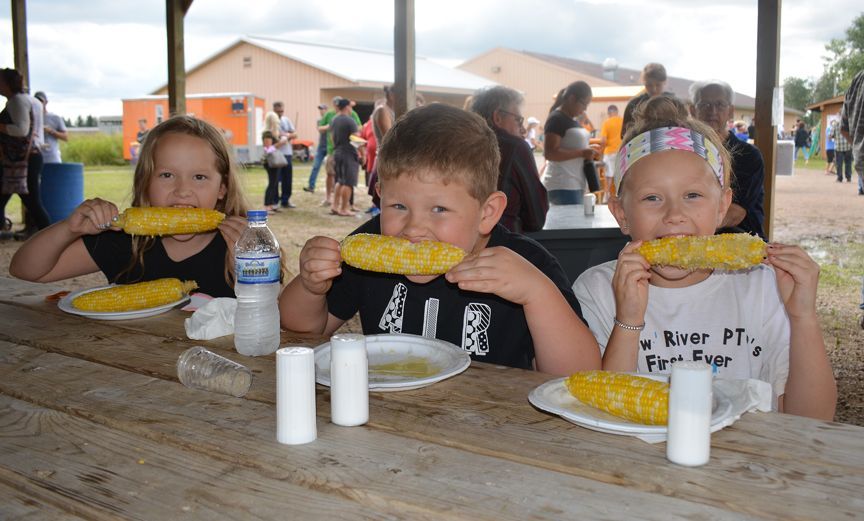 Three children at a picnic table eating corn on the cob outdoors under a covered pavilion