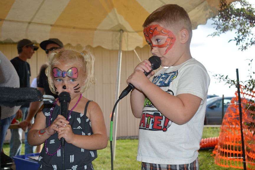 Two children singing into microphones at an outdoor party, wearing face paint and holding toy instruments.