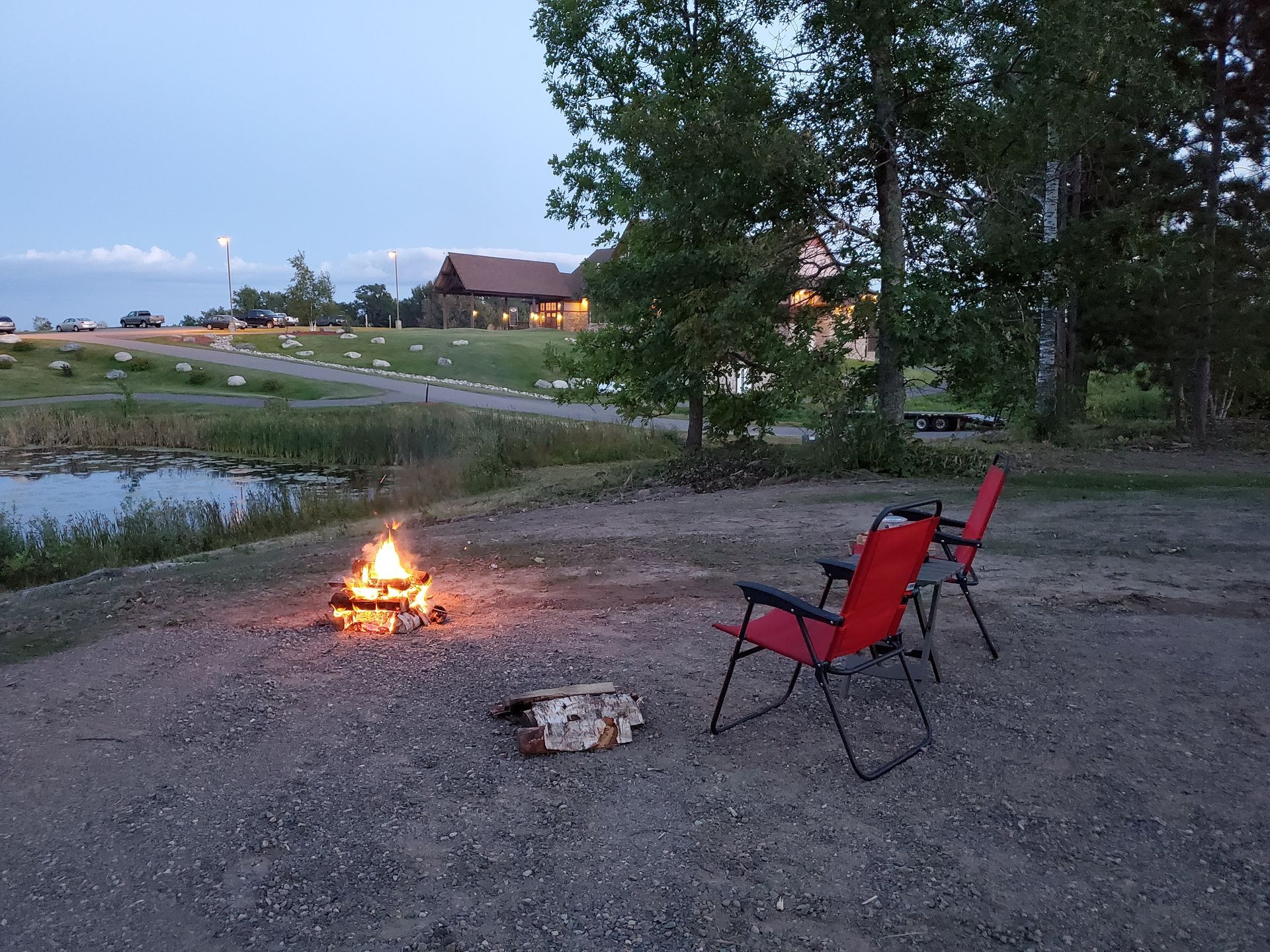 Two red chairs by a campfire on a gravel lakeside at dusk, with trees and cabins in the background