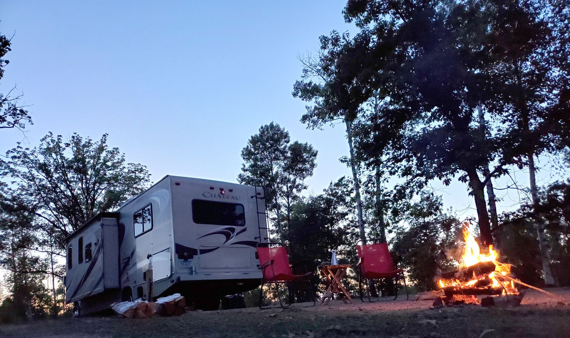 Campsite at dusk with an RV, red chairs, and a campfire among trees