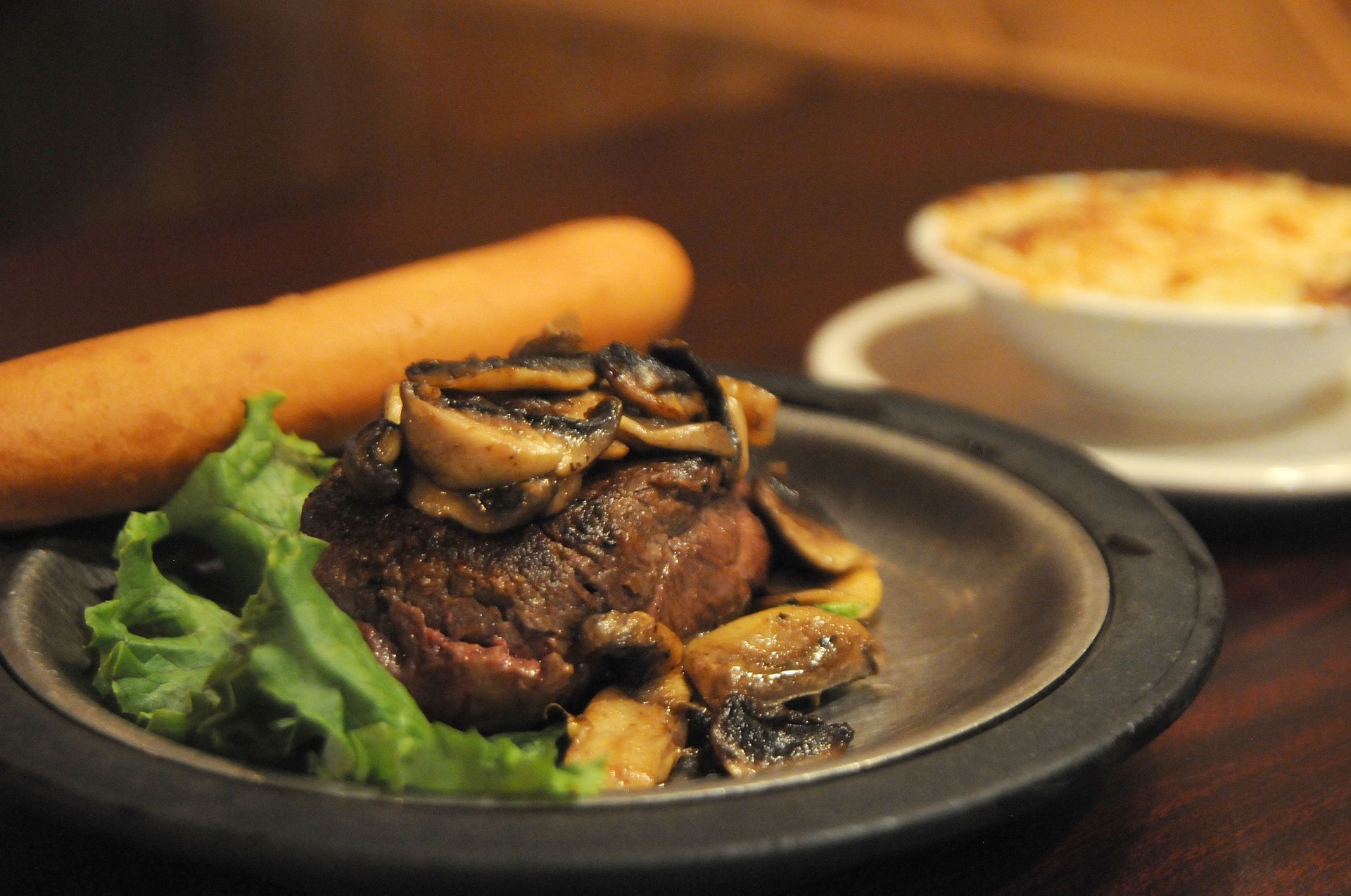 Plated steak topped with mushrooms, served with lettuce, a carrot, and a side dish in the background