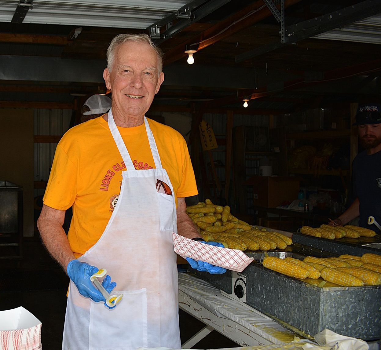 Man in orange shirt and white apron holding a tray of corn in a market stall