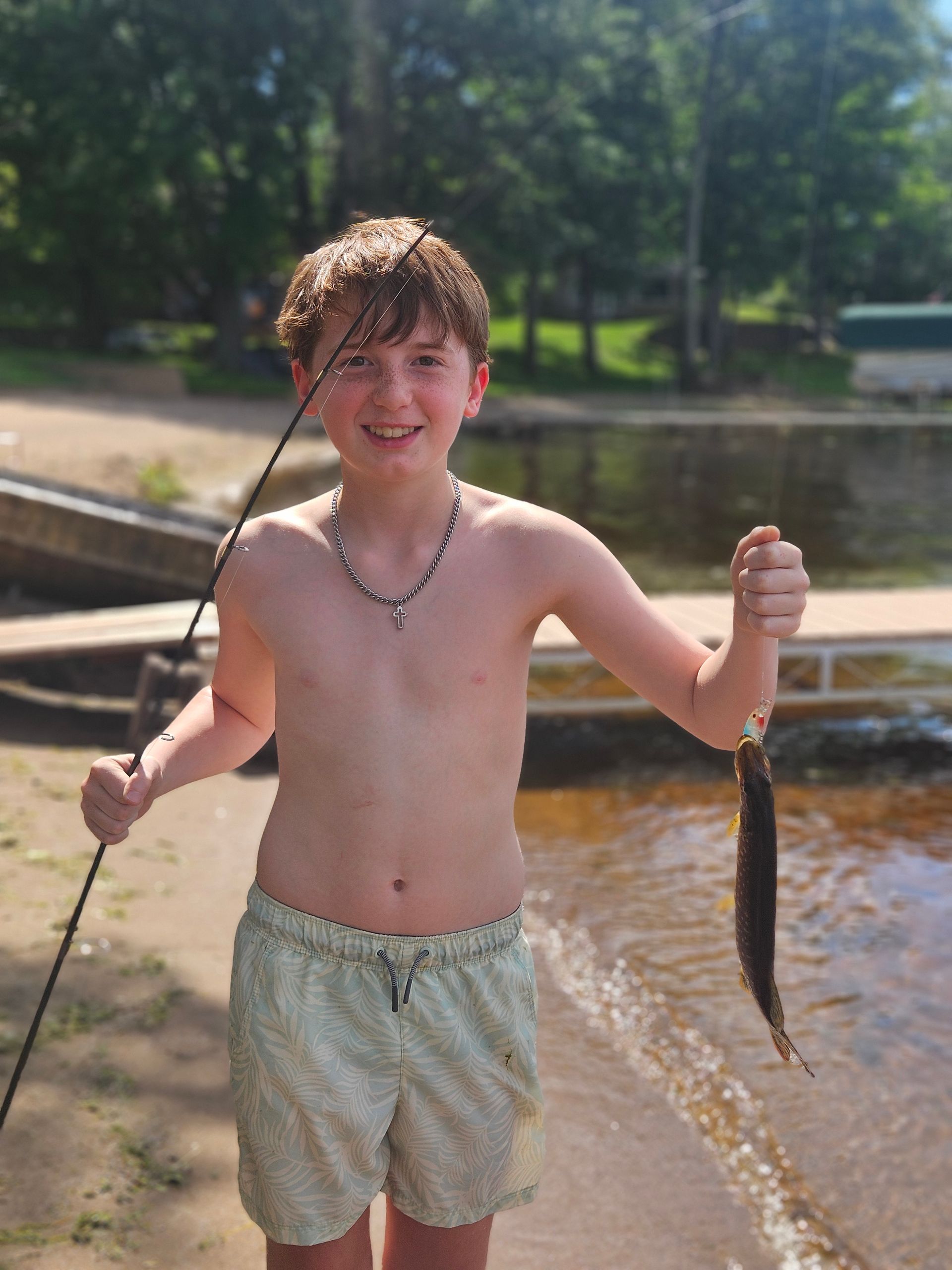 Boy standing in shallow water, holding a fishing line with a fish, smiling at the camera