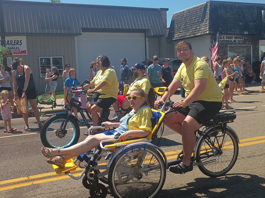 People riding adaptive tricycles in a parade on a sunny street, wearing yellow shirts