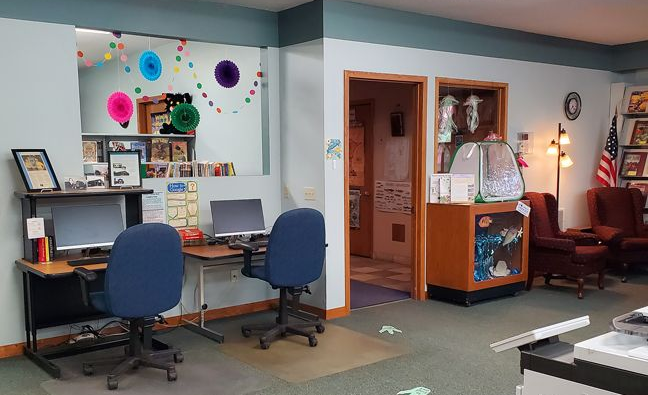 Office with desks, blue chairs, bulletin board, cabinet, and cozy seating area with lamp.