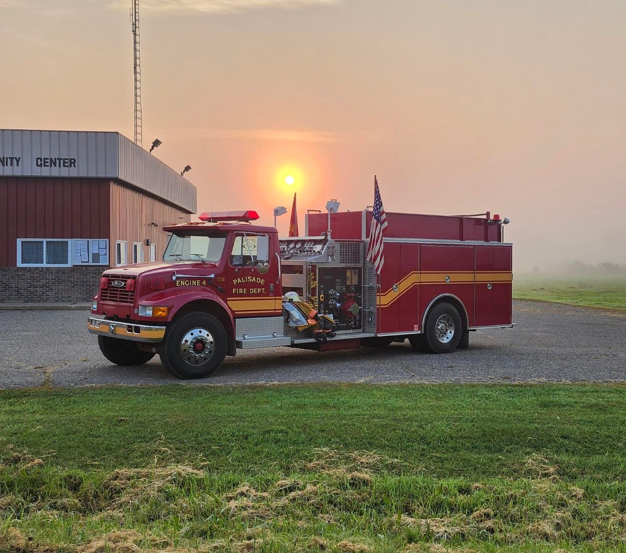 Red fire truck parked on gravel beside a building at sunrise, with a hazy sky and green grass in front.